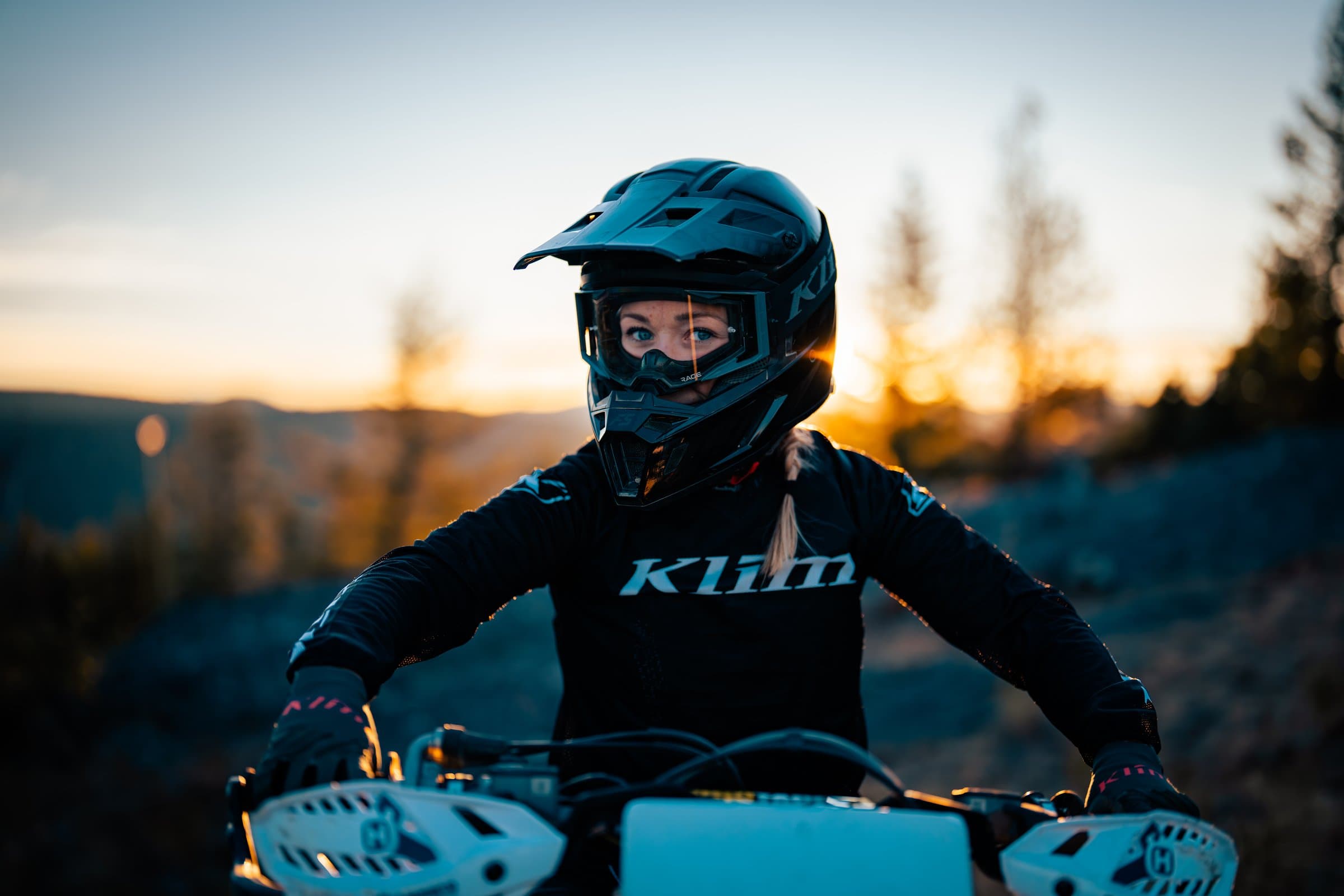 A Dusthunniez rider pauses on a ridge at sunset, golden light behind her, dirt bike in the foreground.